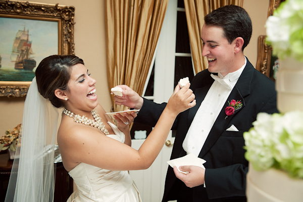 bride and groom feeding each other cake at the reception - bride is wearing ivory dress with full length veil and ivory beaded necklace and groom is wearing black tuxedo with white bowtie and dark red boutonniere - photo by Houston based wedding photographer Adam Nyholt